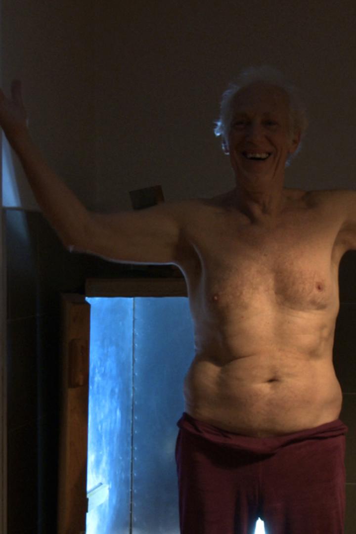 Ein älterer Mann steht mit erhobenen Armen in einem Badezimmer.