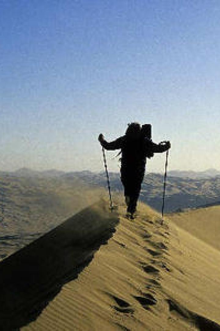 Ein Wanderer mit Rucksack und Stöcken geht über eine Sanddüne.