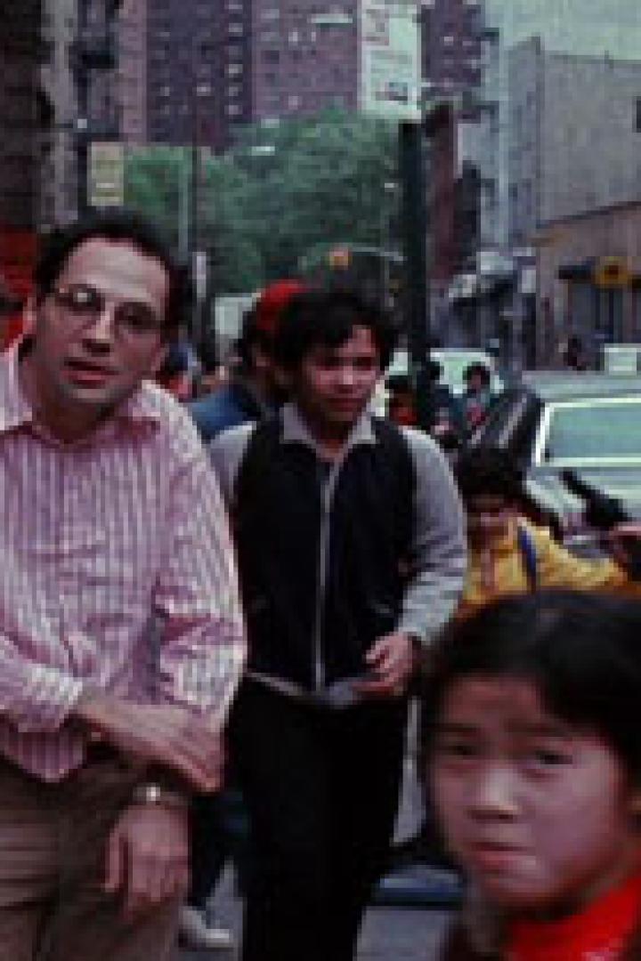 Ein Mann mit Brille und gestreiftem Hemd steht auf einem Bürgersteig in einer belebten Straße.