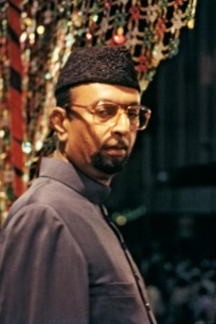 Zwei Männer, einer mit Brille und traditioneller Kopfbedeckung, stehen vor einer Menschenmenge.