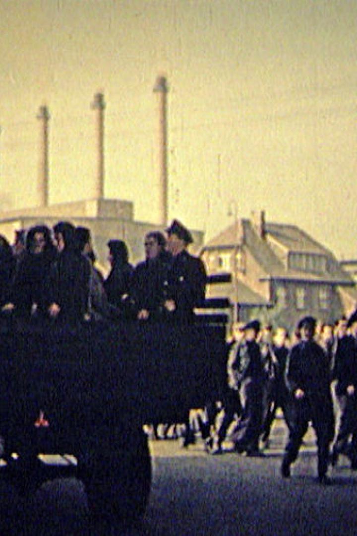 Ein Lastwagen transportiert Menschen in Uniform vor dem Hintergrund einer Industrieanlage.