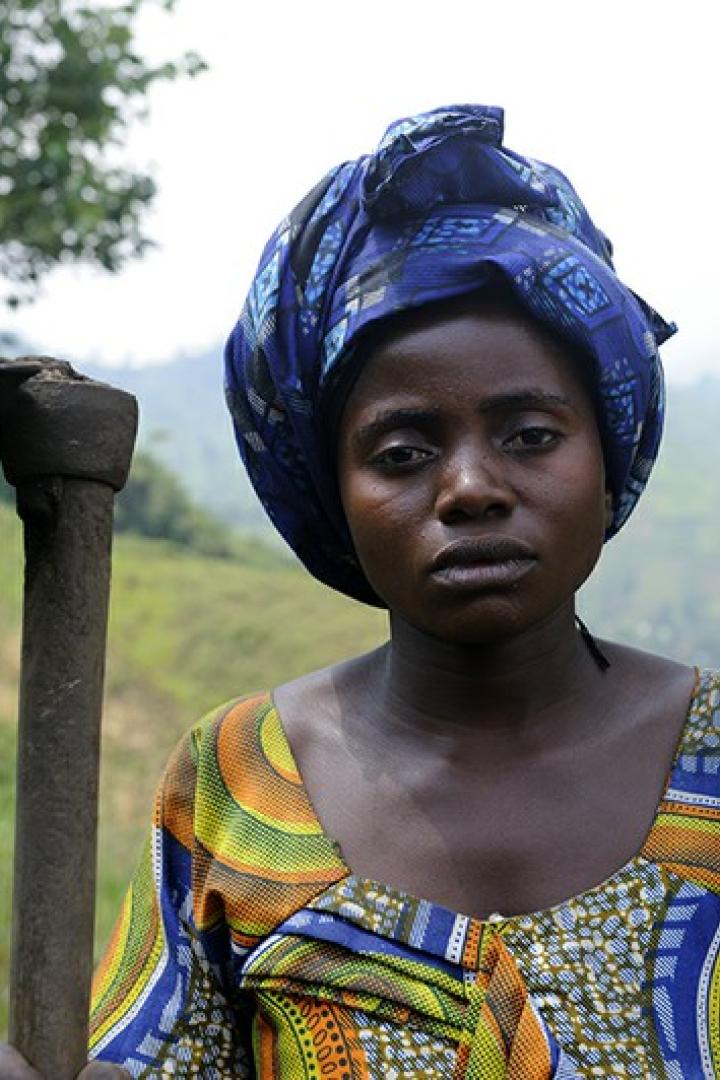 Eine Frau mit Kopftuch hält eine Hacke vor einer hügeligen Landschaft.
