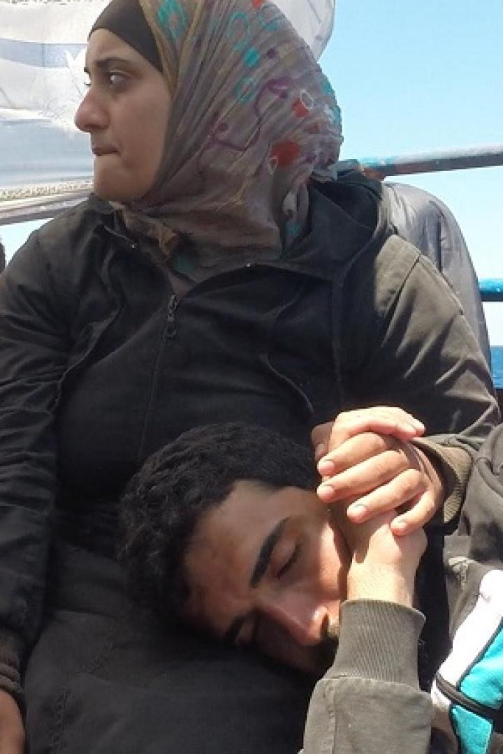 Eine Frau mit Kopftuch hält die Hand eines schlafenden Mannes auf einem Boot.