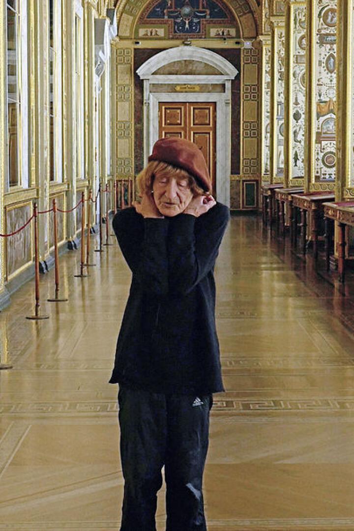 Eine ältere Frau mit Hut steht in einem reich verzierten Korridor.