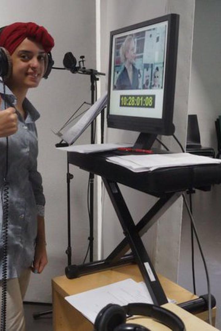 Eine Frau mit Kopfbedeckung und Kopfhörern steht vor einem Monitor in einem Studio und gibt einen Daumen hoch.