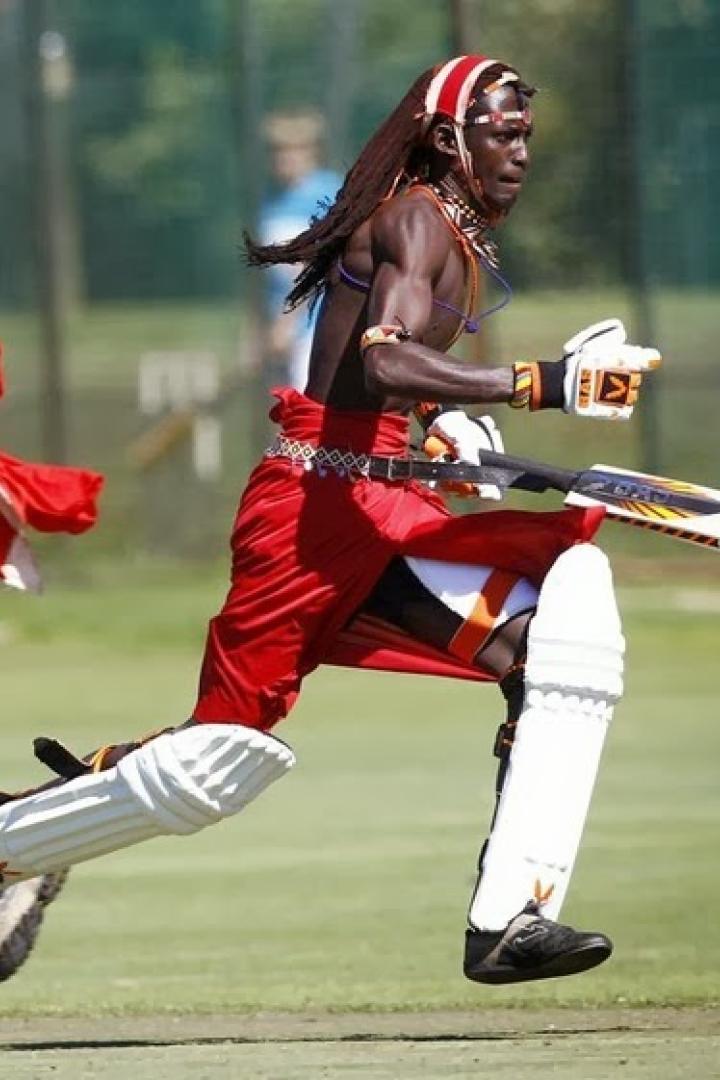 Zwei kenianische Cricketspieler in traditioneller Kleidung rennen über ein grünes Feld.