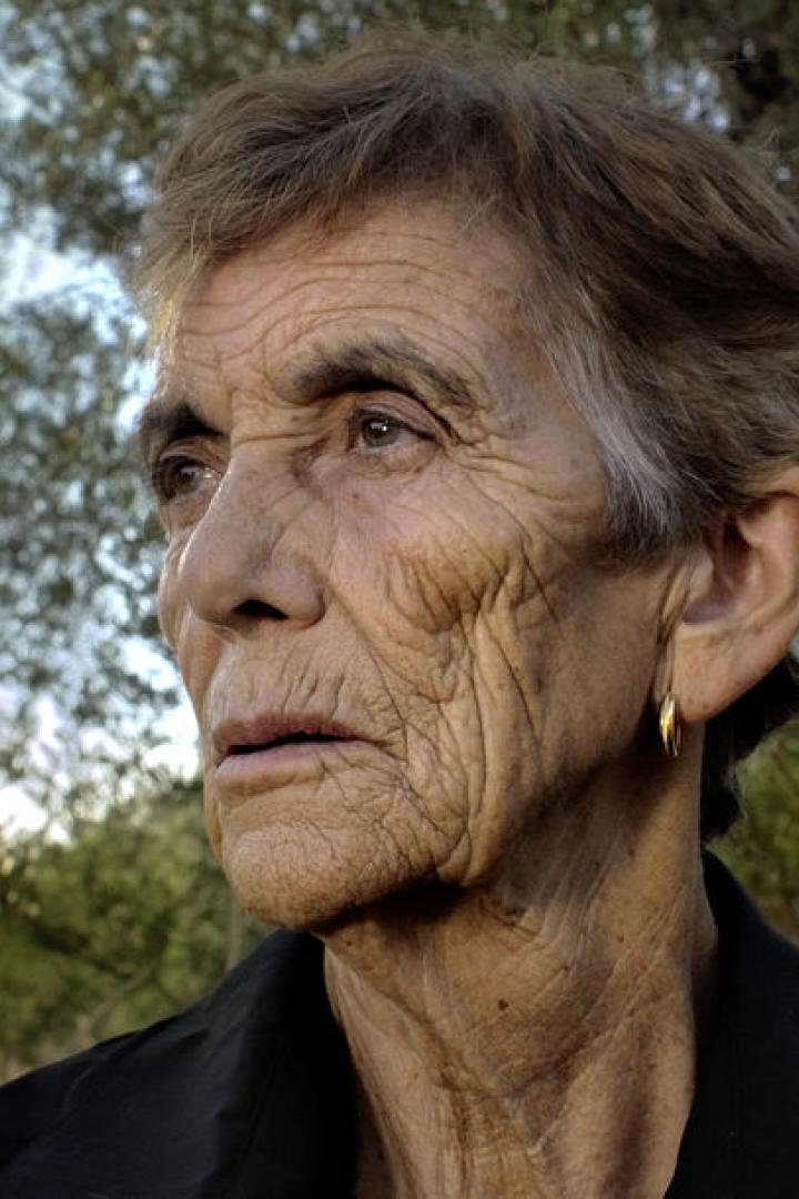 Porträt einer älteren Frau mit kurzem Haar und Ohrring vor Bäumen.