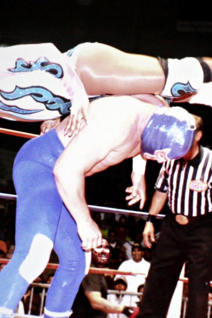 Eine Wrestlerin führt einen Move gegen einen maskierten Gegner im Ring aus, während ein Schiedsrichter zusieht.