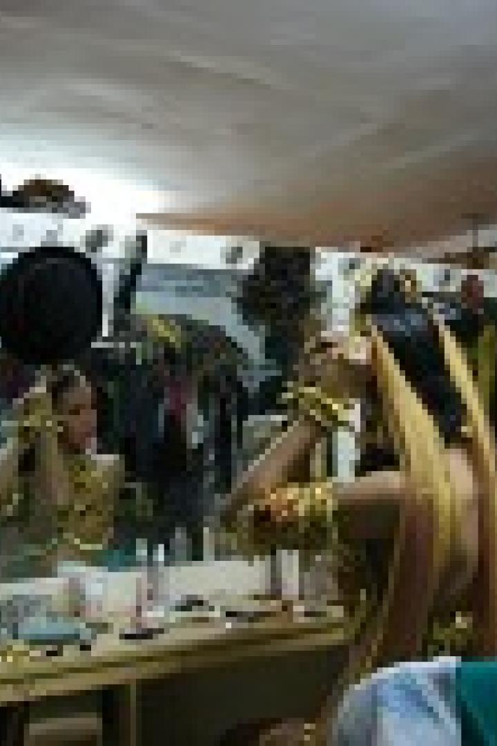 Eine Dragqueen schminkt sich vor einem Spiegel, umgeben von Kostümen und Accessoires.