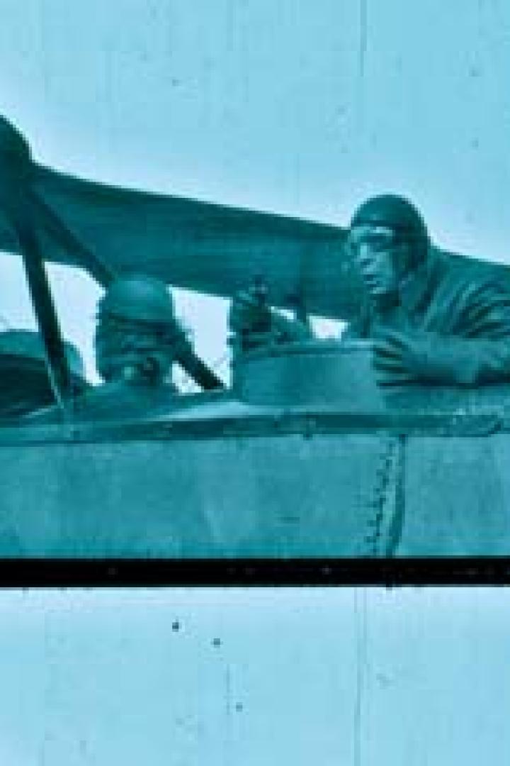 Ein Pilot und zwei weitere Personen sitzen in einem alten Doppeldecker-Flugzeug.