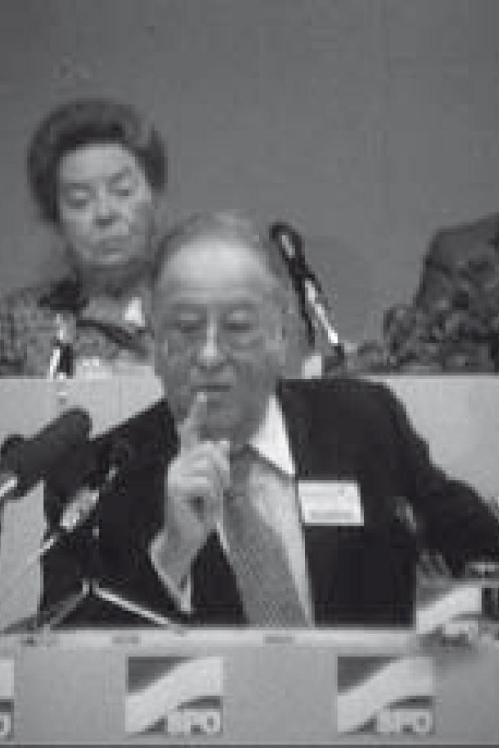 Ein Mann hält eine Rede vor Mikrofonen, während zwei Personen im Hintergrund sitzen.