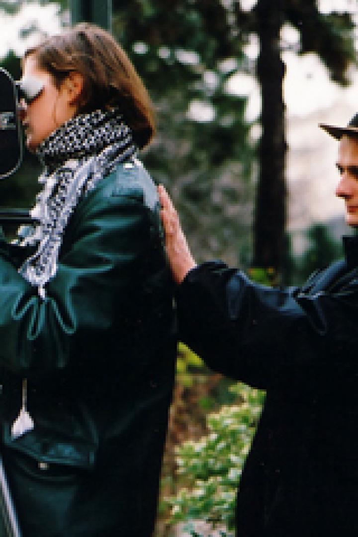 Eine Frau filmt mit einer alten Filmkamera, während ein Mann ihr die Hand auf den Rücken legt.