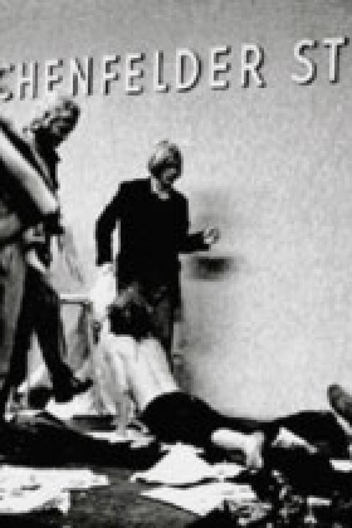 Eine Schwarzweißaufnahme von Menschen, die vor einer Wand mit der Aufschrift „Münchenfelder Strasse“ stehen.