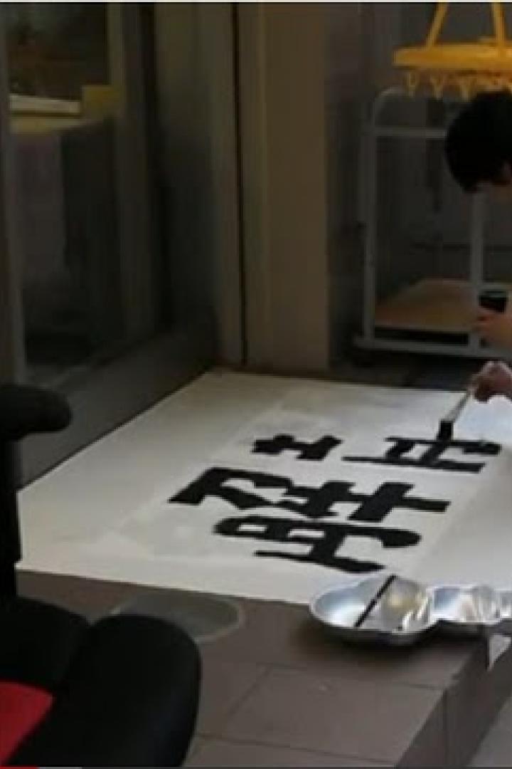 Ein Mann kniet und malt chinesische Schriftzeichen auf ein großes Blatt Papier.
