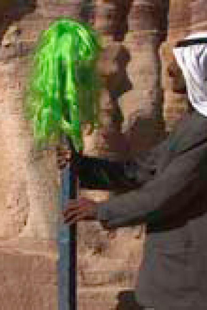 Ein Mann in traditioneller arabischer Kleidung hält eine grüne Perücke vor einer Felswand.