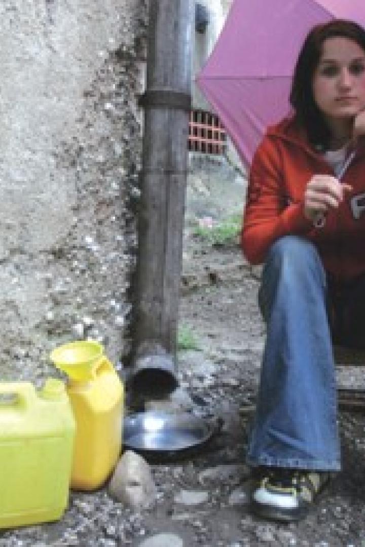 Eine junge Frau sitzt mit einem rosa Regenschirm neben einem Fallrohr und gelben Kanistern.