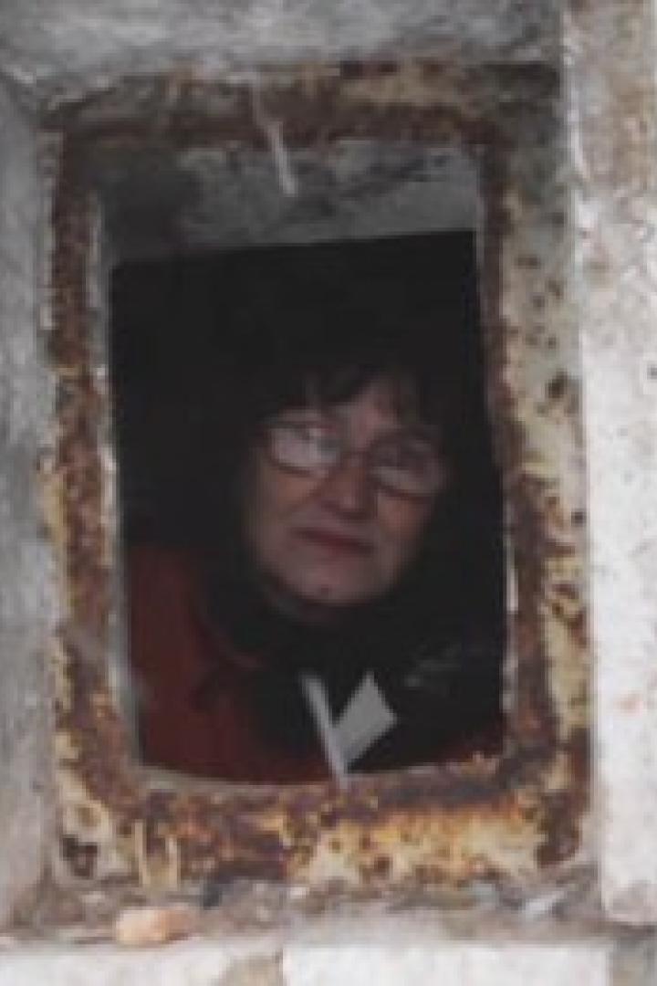 Eine Frau mit Brille schaut aus einer kleinen, rechteckigen Öffnung in einer Mauer.