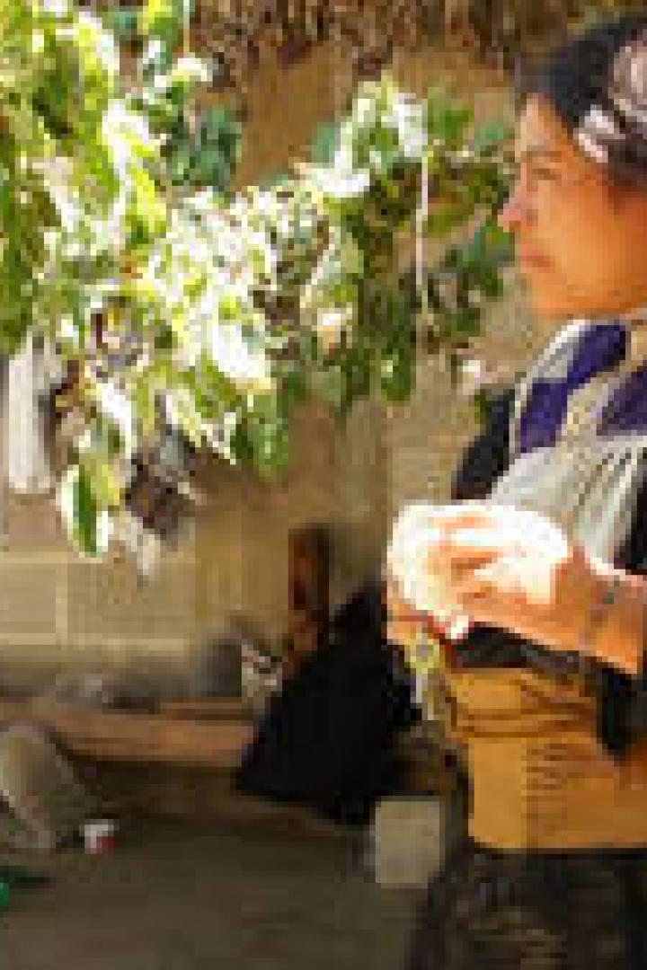 Eine indigene Frau steht vor einem einfachen Haus und blickt nach rechts.