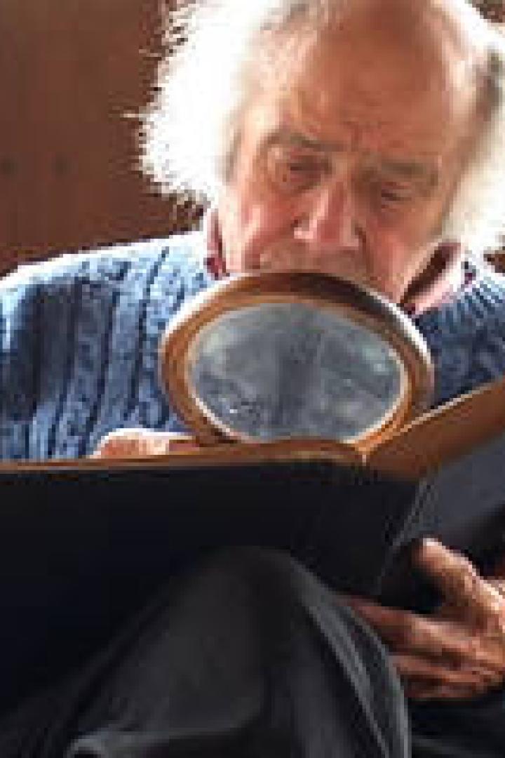 Ein älterer Mann mit weißem Haar betrachtet ein Buch durch eine Lupe.