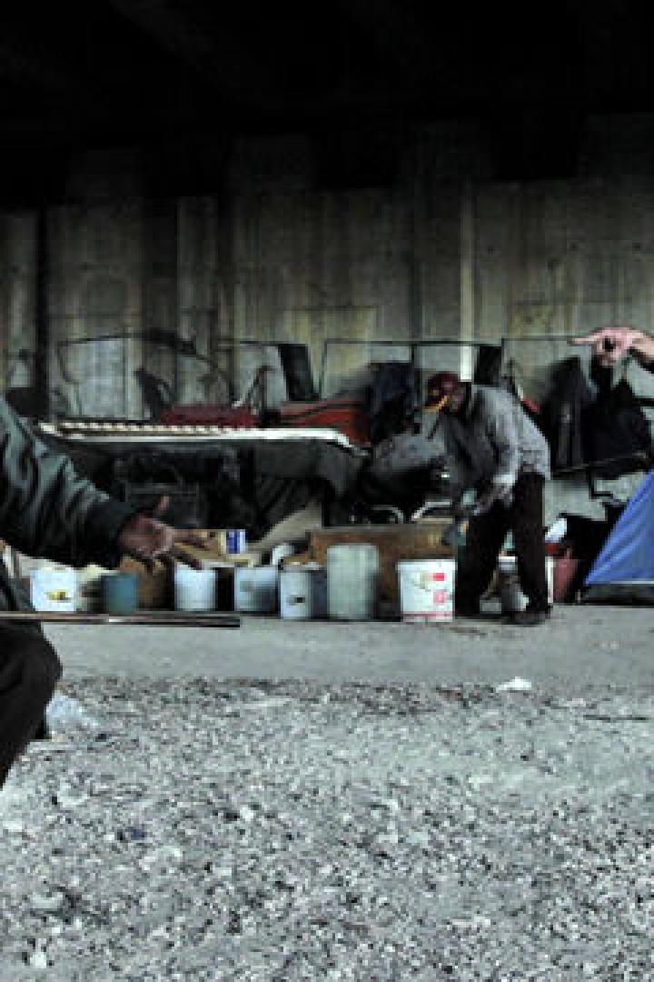 Unter einer Brücke sitzen und stehen mehrere Menschen in einem improvisierten Lager.