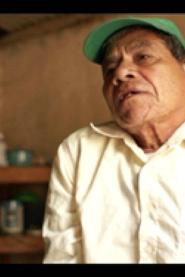 Ein älterer Mann mit grüner Kappe spricht in einem einfachen Raum.