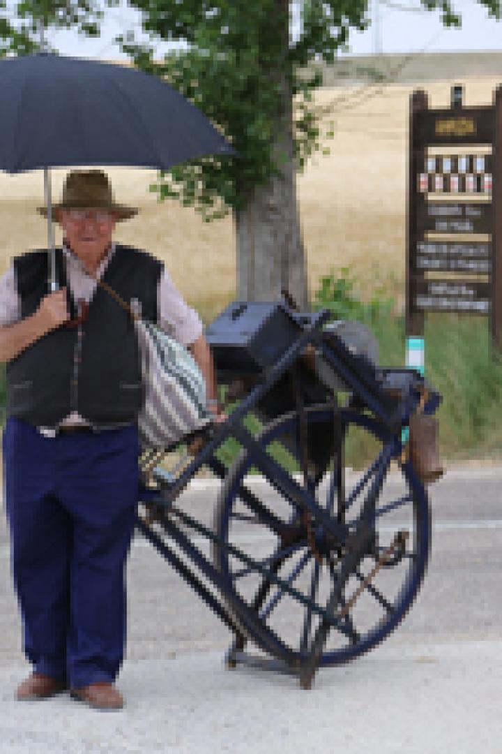 Ein älterer Mann mit Hut und Regenschirm steht neben einem beladenen Karren am Straßenrand.