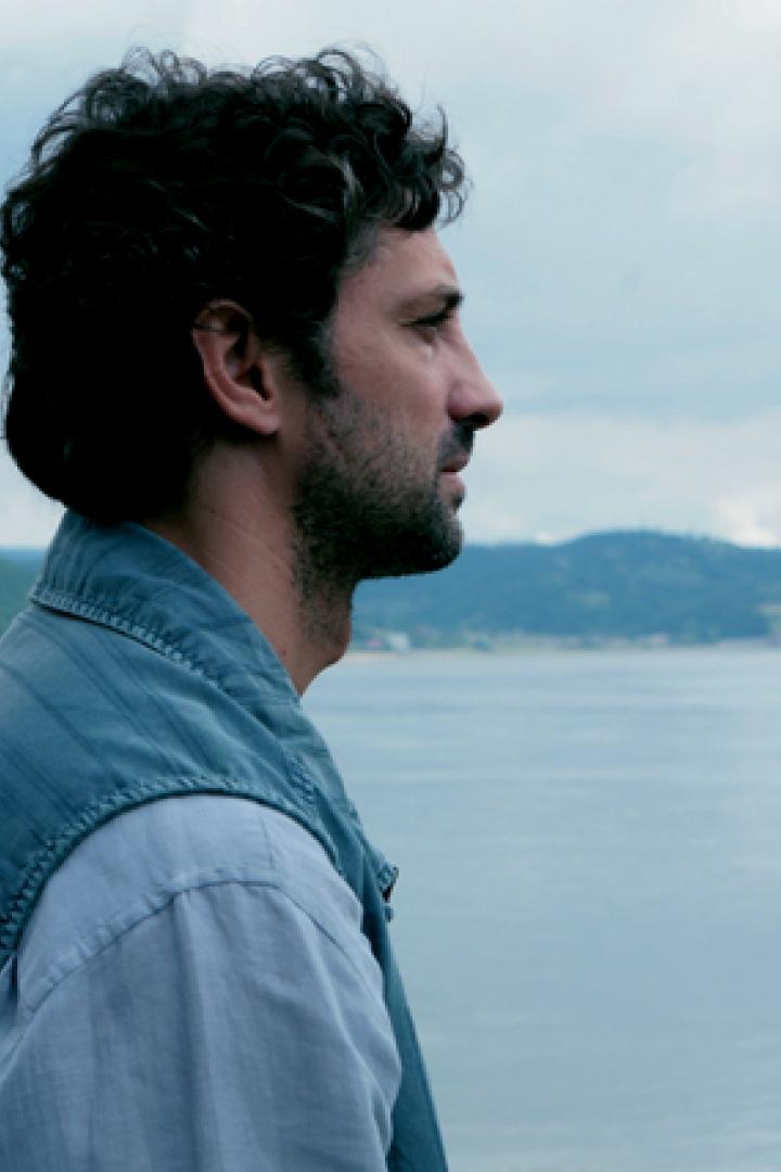 Ein Mann blickt auf einen ruhigen See, umgeben von bewaldeten Hügeln.