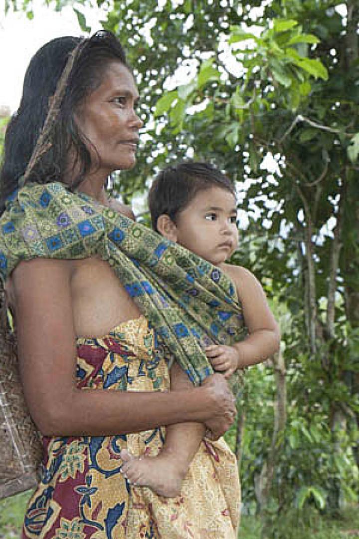 Eine Frau trägt ihr Kind in einer Tragevorrichtung durch eine ländliche Gegend.