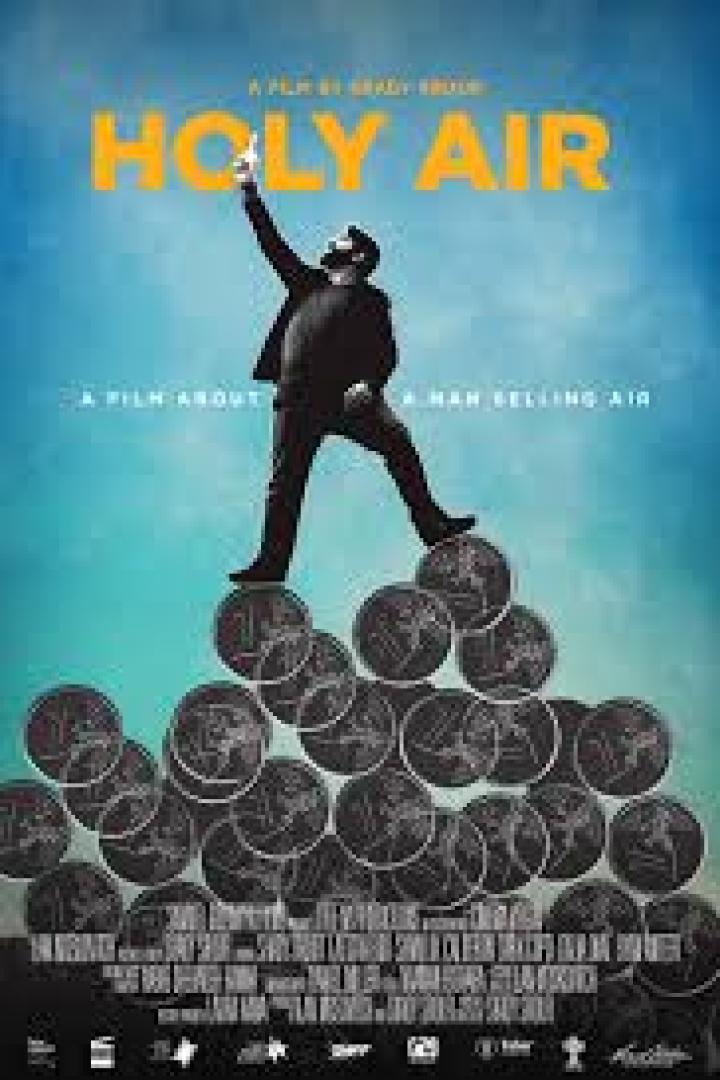 Ein Mann steht auf einem Haufen Münzen vor dem Filmplakat von „Holy Air“.