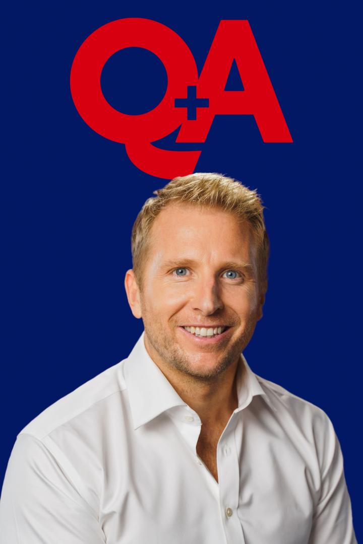 Ein lächelnder Mann mit blonden Haaren vor einem blauen Hintergrund mit dem roten Schriftzug „QA“.