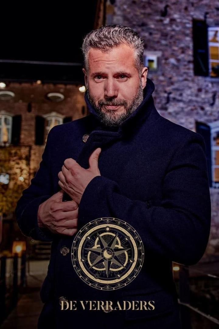 Ein Mann in einem dunklen Mantel vor einem Backsteingebäude, mit dem Logo von „De Verraders“.