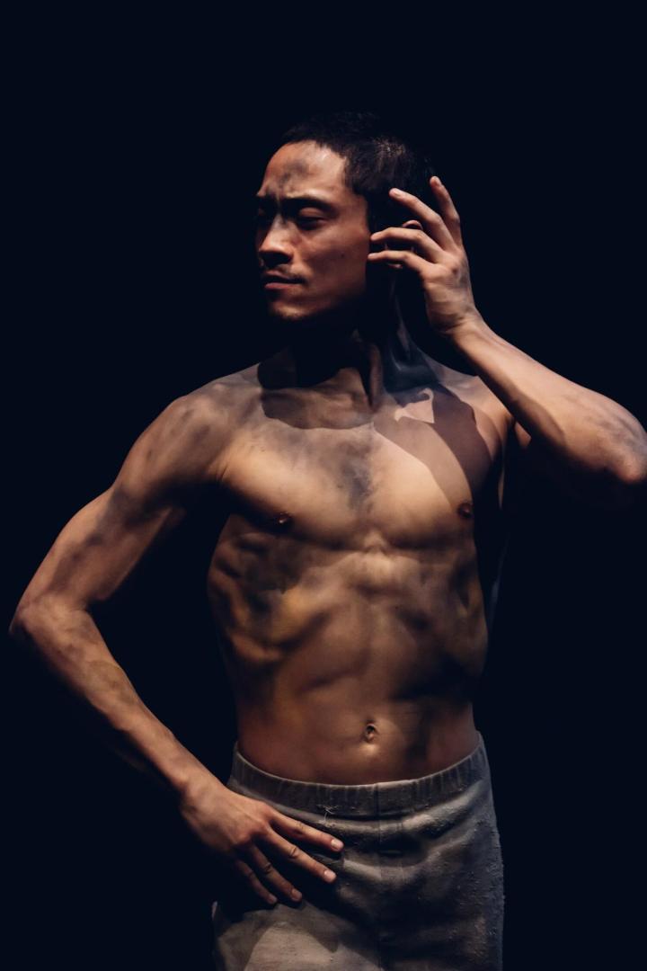 Ein Mann mit nacktem Oberkörper und schmutziger Haut posiert vor einem dunklen Hintergrund.