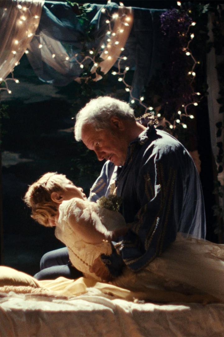 Ein älterer Mann hält ein junges Mädchen im Arm, beide in festlicher Kleidung.