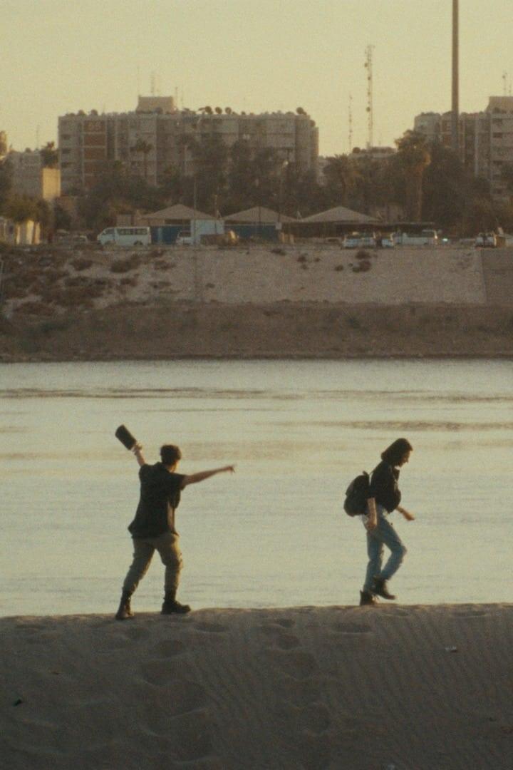 Zwei Personen spazieren an einem Flussufer entlang, eine wirft einen Gegenstand ins Wasser.