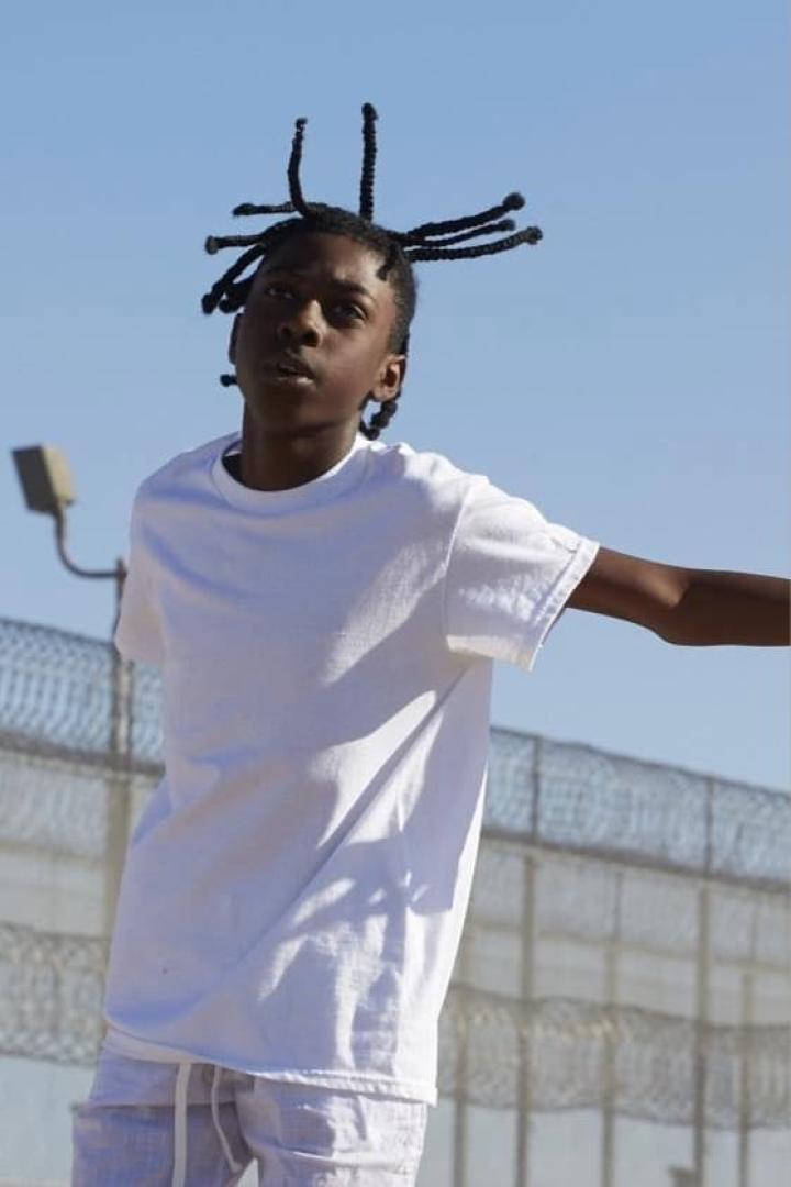 Ein junger Mann mit geflochtenen Zöpfen trägt ein weißes T-Shirt vor einem Zaun.