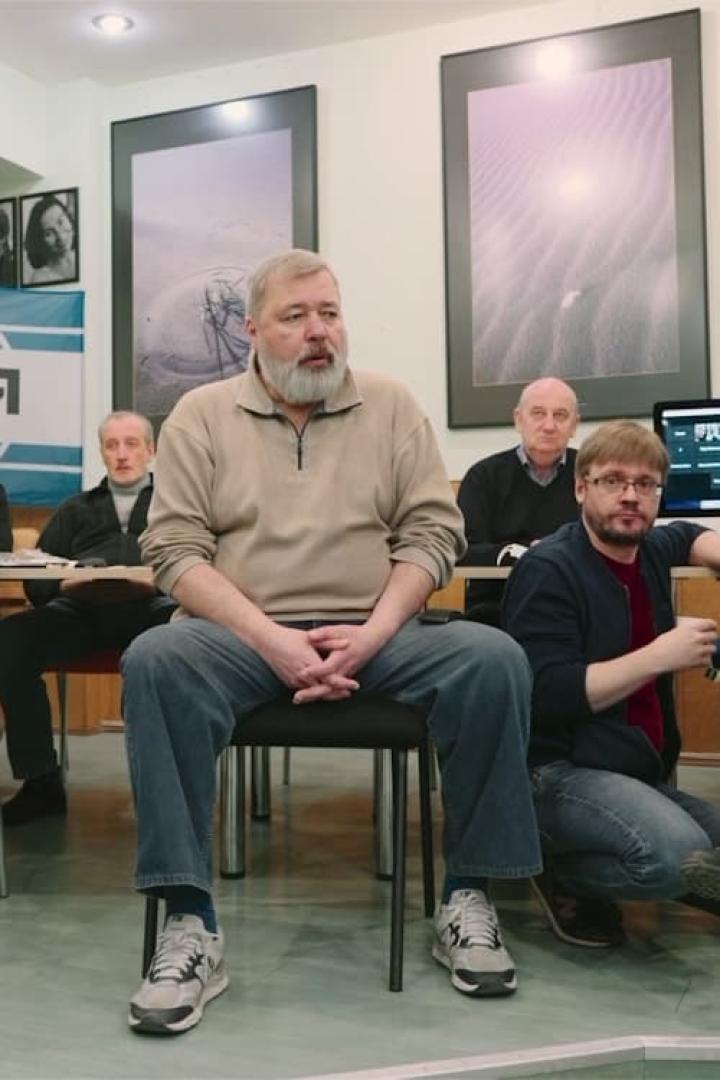 Ein Mann mit Bart sitzt auf einem Stuhl vor anderen Personen.