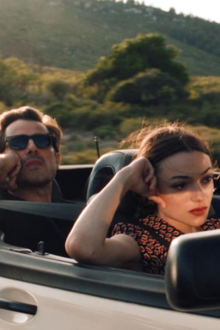 Ein Mann mit Sonnenbrille und eine Frau fahren in einem Cabriolet durch eine grüne Landschaft.