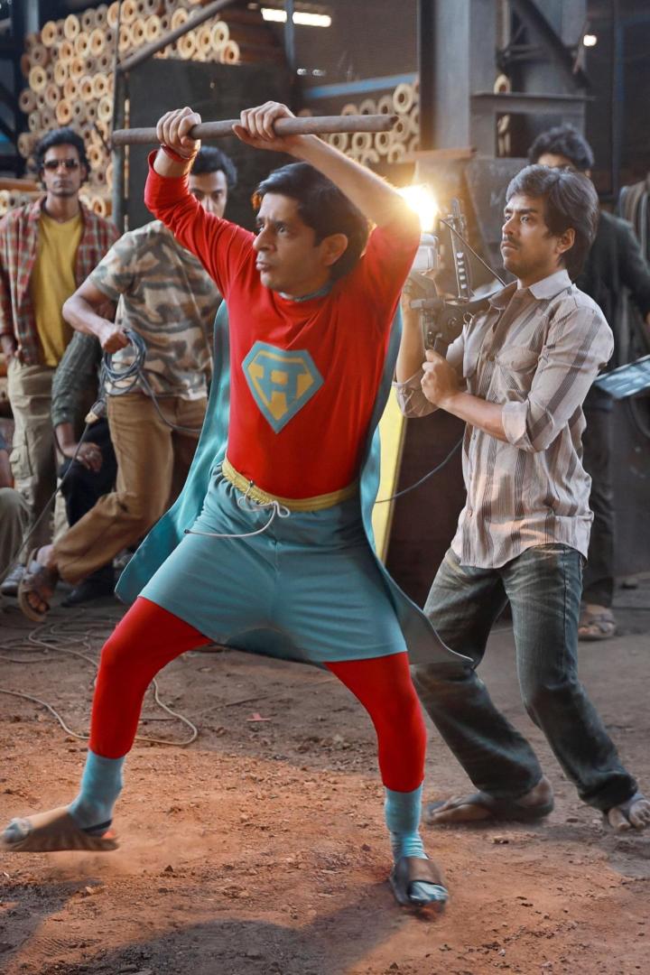 Ein Mann in einem Superheldenkostüm schwingt eine Stange vor einer Filmcrew.