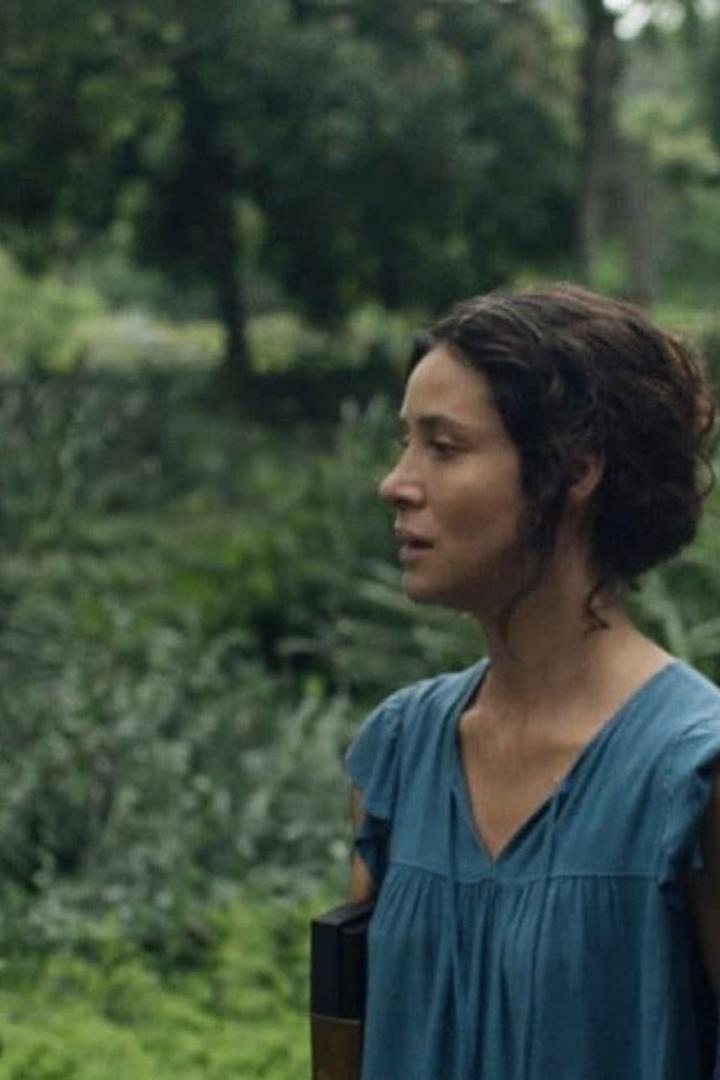 Eine Frau mit dunklen Haaren und blauem Kleid steht vor einer grünen Landschaft.