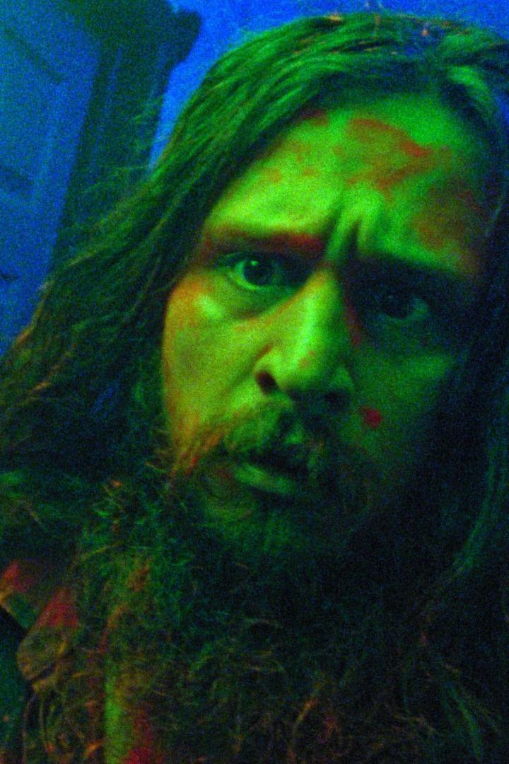 Ein Mann mit Bart und langen Haaren, beleuchtet in grünem Licht, blickt in die Kamera.