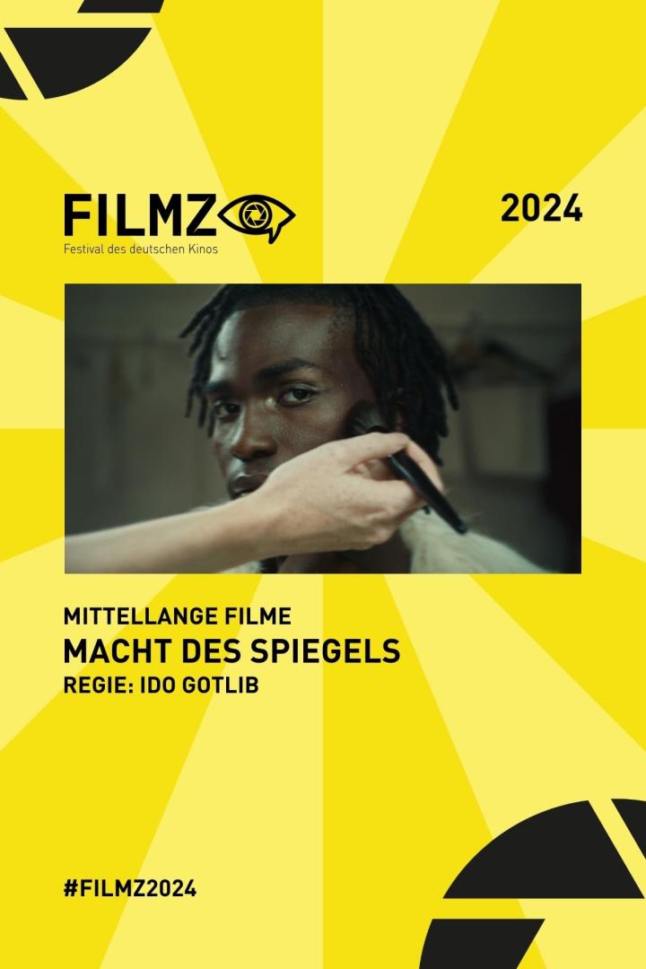 Einem Mann wird Make-up aufgetragen, im Hintergrund das Filmplakat „Macht des Spiegels“.