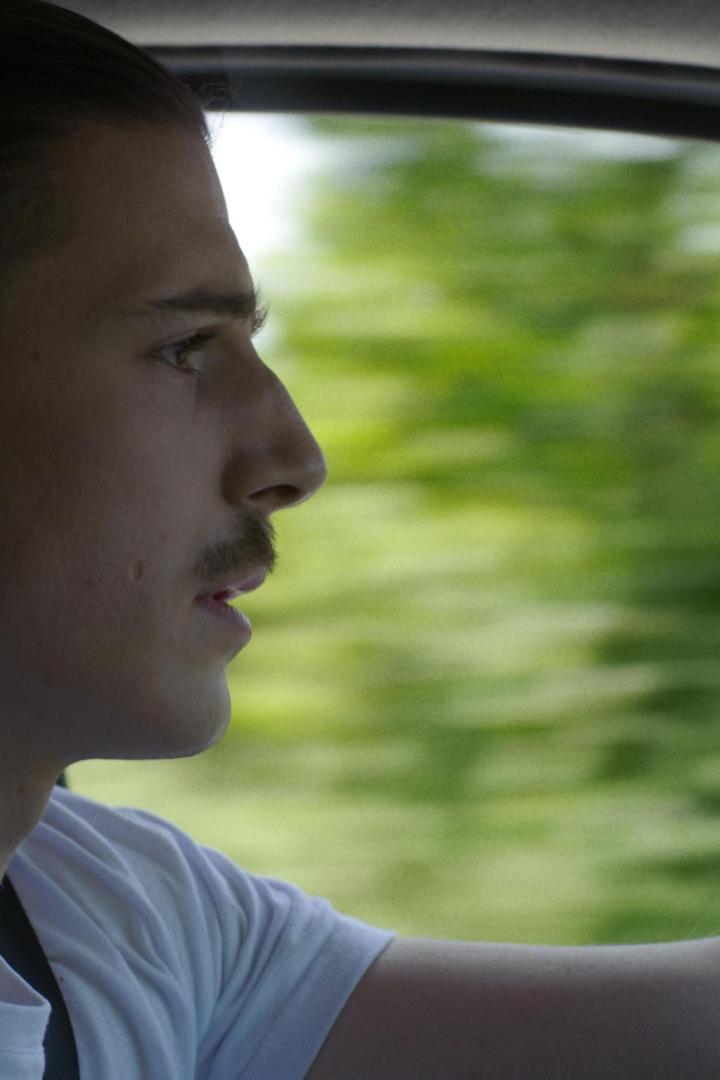 Ein Mann mit Schnurrbart fährt Auto; im Hintergrund verschwimmt die grüne Landschaft.