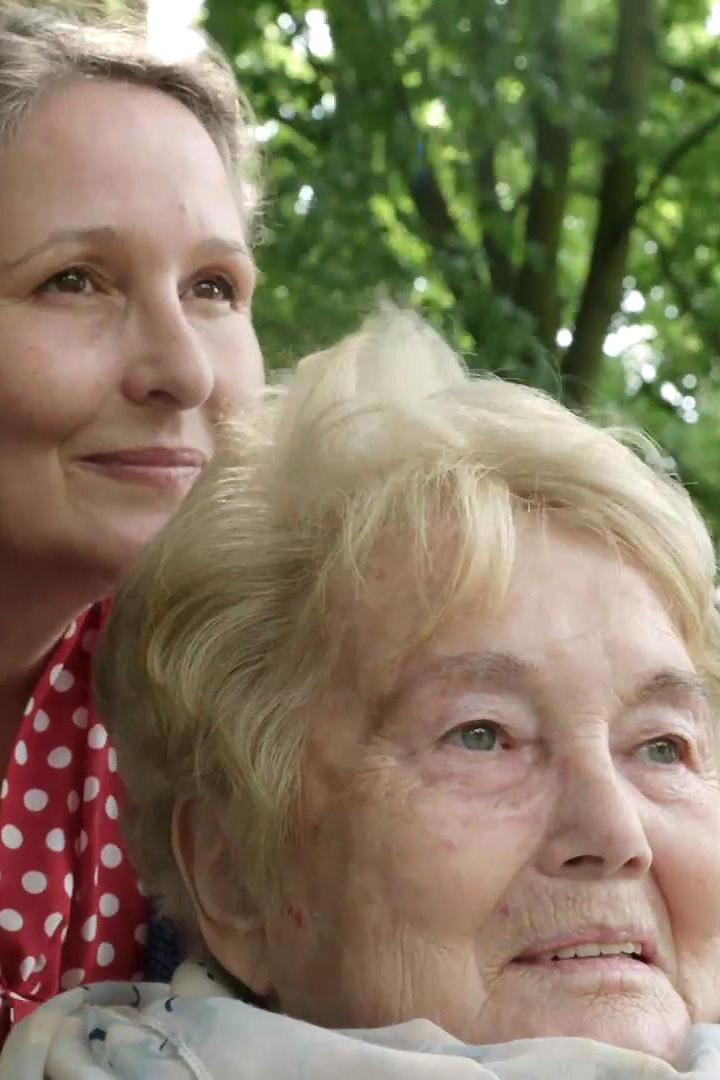 Eine ältere Frau mit blonden Haaren und eine jüngere Frau im gepunkteten Kleid schauen nach oben.