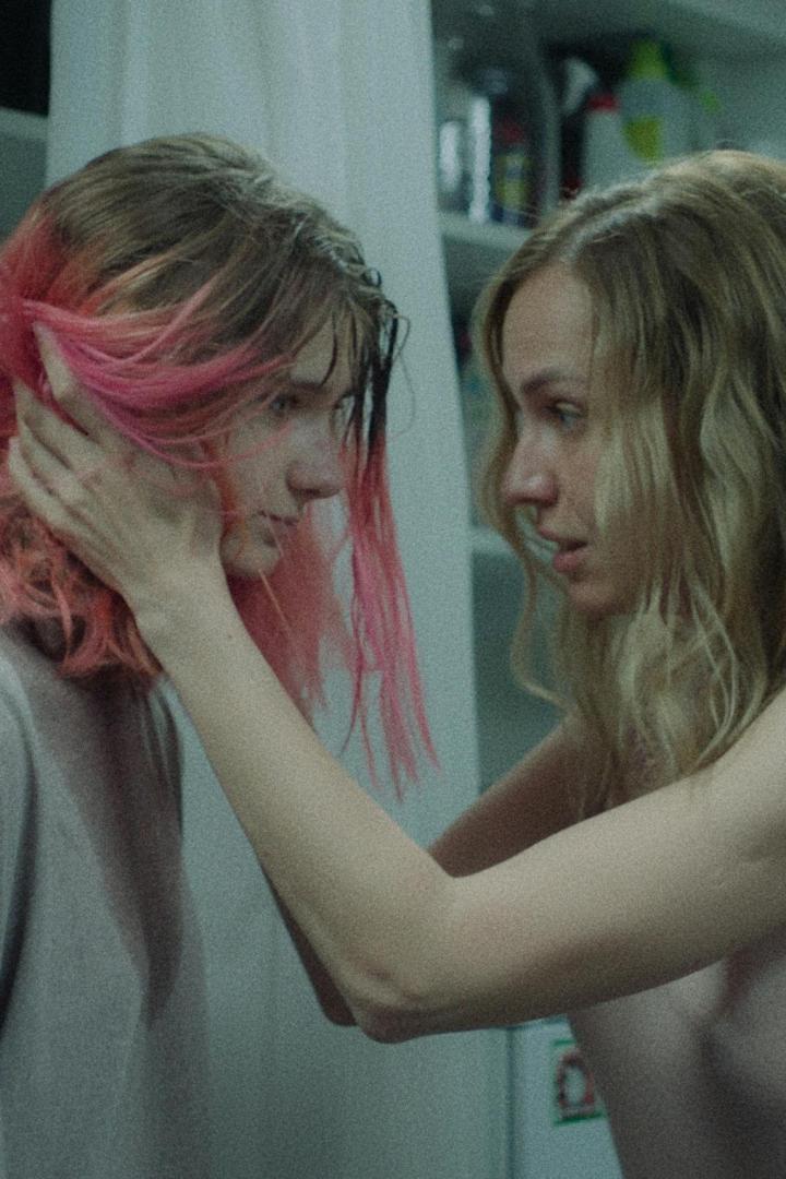 Eine Frau mit blonden Haaren berührt das Gesicht einer anderen Frau mit pink gefärbten Haaren.