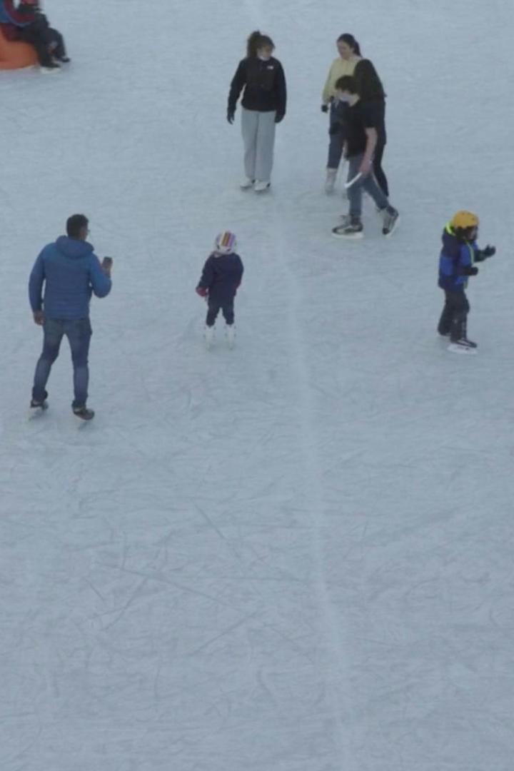 Mehrere Personen, darunter Kinder mit Helmen, laufen auf einer Eisbahn Schlittschuh.