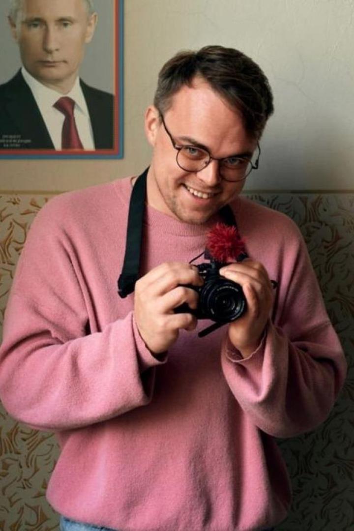 Ein Mann mit Brille und rosa Pullover hält eine Kamera in den Händen. Im Hintergrund hängt ein Porträt von Putin.