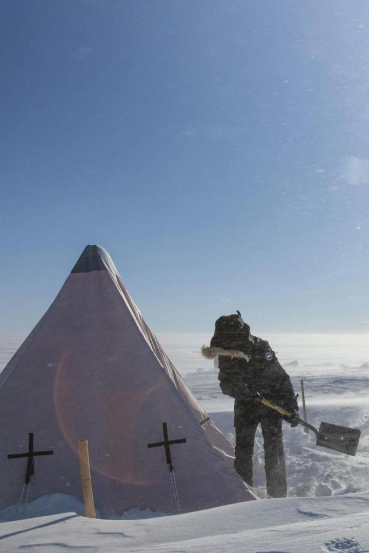 Eine Person schaufelt Schnee vor einem Zelt in einer verschneiten Landschaft.