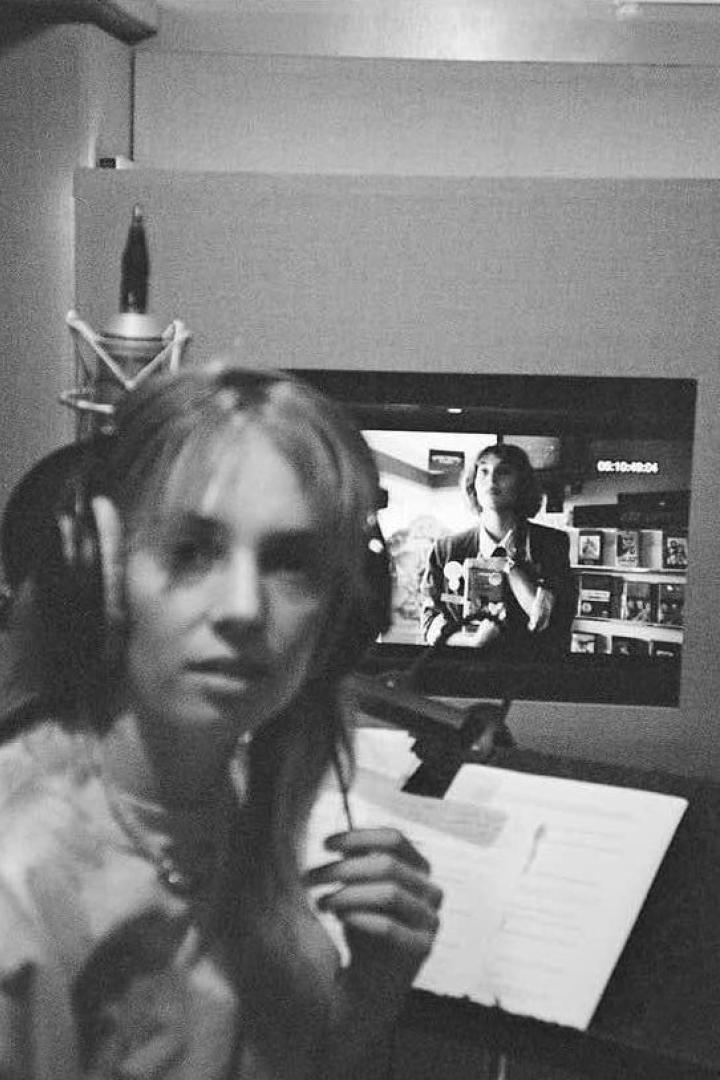 Eine Frau mit Kopfhörern vor einem Mikrofon, im Hintergrund ein Monitor mit einer Filmszene.