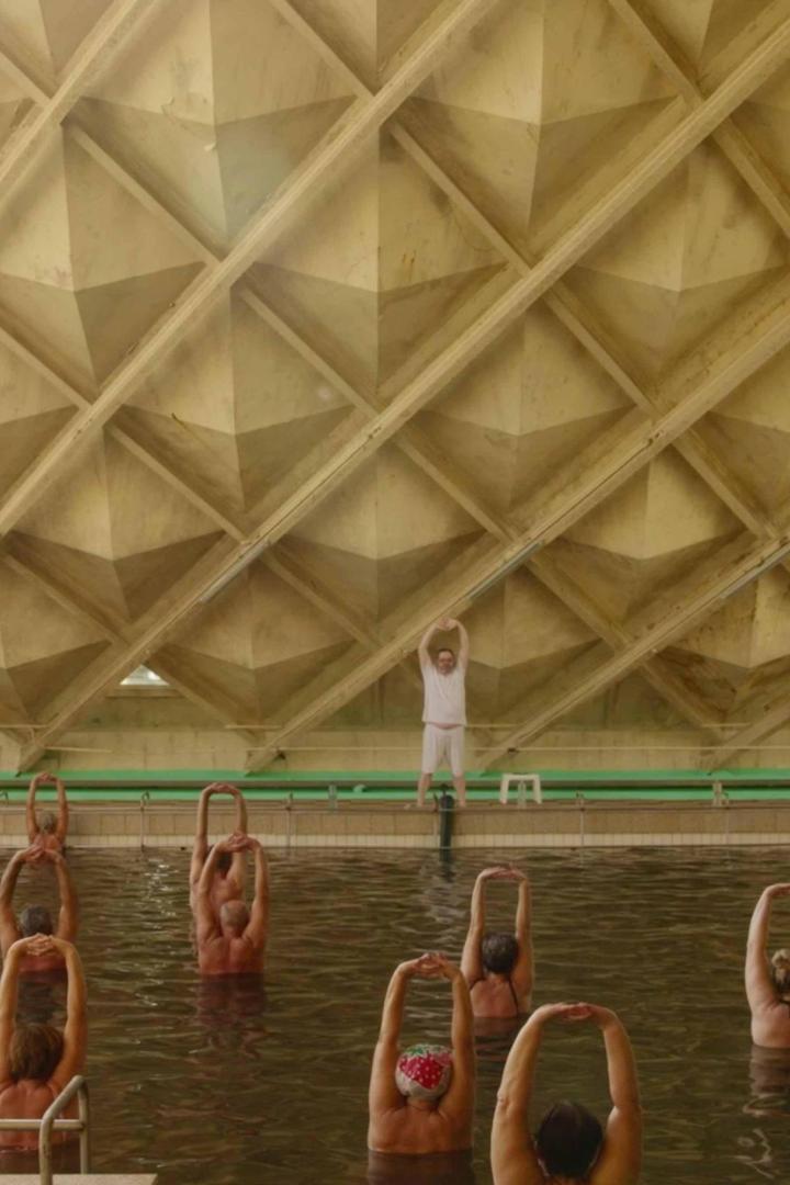 Eine Gruppe von Menschen macht Wassergymnastik in einem Schwimmbad mit markanter Deckenstruktur.