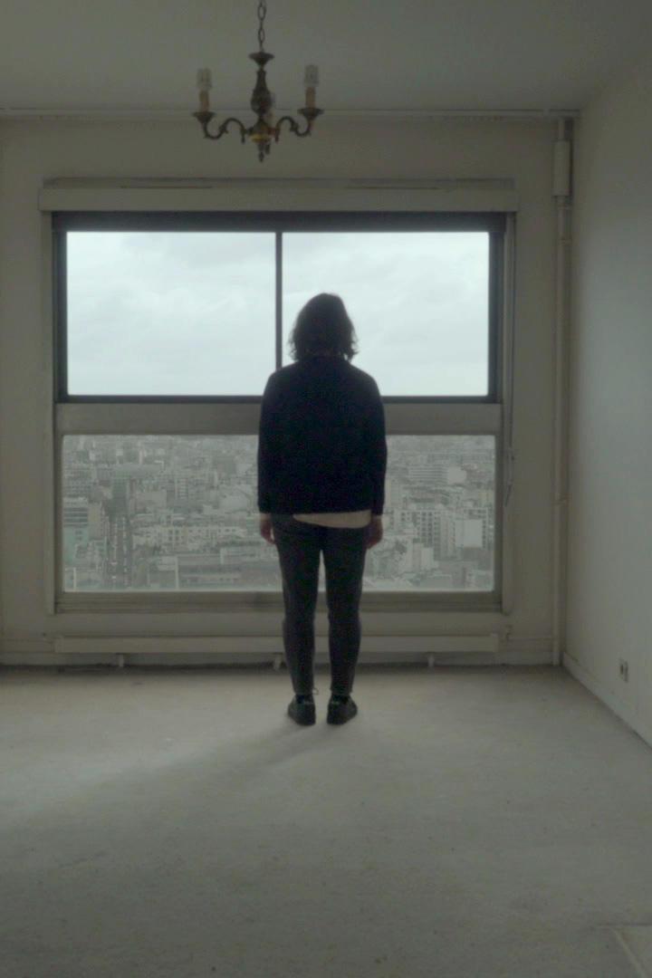 Eine Person steht in einem leeren Raum und blickt aus dem Fenster auf die Stadt.